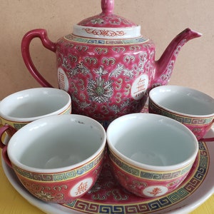 May include: A pink and white porcelain tea set with a teapot and four cups. The teapot features a floral design and the cups have a geometric design. The set is on a white plate with a red and blue border.
