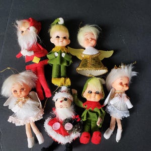 May include: A collection of seven vintage-style Christmas ornaments featuring dolls dressed as Santa Claus, elves, and angels. The figures have painted facial features, synthetic hair, and fabric clothing in red, green, white, and gold metallic tones.