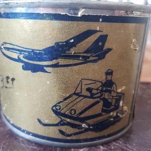 May include: Vintage metal container with a gold background and blue illustrations. The illustrations include an airplane and a snowmobile with a driver. The container has some wear and tear, adding to its vintage appeal.