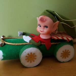 Peut inclure: Une voiture jouet verte avec une roue blanche et dorée, un colis rayé rouge et blanc à l'arrière et un poupon lutin souriant au volant.
