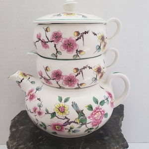 May include: A white ceramic teapot with a floral design. The teapot is stacked on top of two smaller teacups with matching floral designs. The teapot and teacups have a green trim around the rim. The teapot has a bird and flowers on the side. The teapot is sitting on a grey rock.
