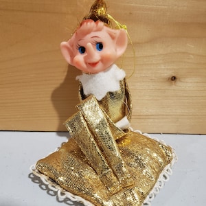 May include: A gold glitter Christmas elf ornament sitting on a gold glitter pillow. The elf has a white fur collar and blue eyes.
