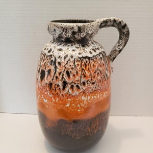 May include: A brown, orange, and white ceramic vase with a speckled glaze. The vase has a rounded body and a handle.