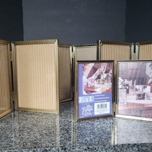 Peut inclure: Un ensemble de cadres photo pliants à plusieurs panneaux avec une finition dorée métallique et des bordures fines et texturées. Les cadres présentent des inserts beiges à nervures verticales. Une étiquette indique 9 x 13 cm et Royal.