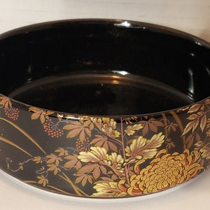 May include: A black ceramic bowl with a gold and brown floral design. The bowl has a wide rim and a rounded bottom.