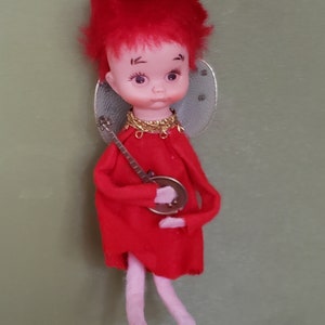 May include: A red felt angel ornament with a gold crown, wings, and a necklace. The angel is holding a small banjo and has a pink face with painted black eyes and eyebrows.