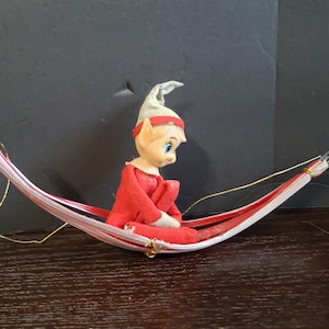 May include: A small elf figurine with a pale face, blue eyes, and a pointed white hat, wearing a red felt outfit. The elf is sitting inside a miniature hammock constructed from thin, flexible red and white striped plastic strips secured with gold wire.