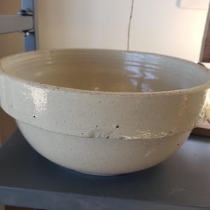 Peut inclure: Un grand bol mélangeur en céramique blanc cassé, de forme ronde, avec une finition brillante et tachetée. Le bol présente une crête horizontale marquée sur l'extérieur supérieur, des petites indentations sur le bord et un éclat visible.