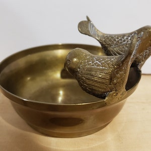 May include: A brass bowl with two bird-shaped handles. The bowl is round and has a smooth, shiny surface. The birds are facing each other and have detailed features.
