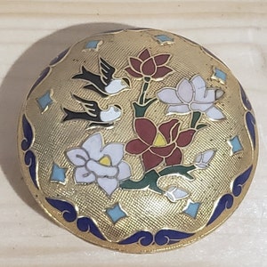 May include: A round, gold-coloured brooch with a blue and white enamel design. The design features three birds, a red flower, and two white flowers.