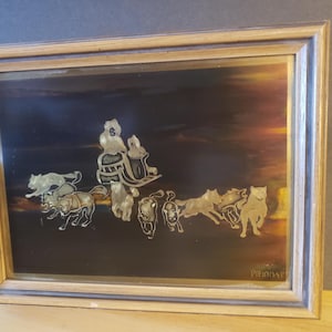 May include: A framed artwork depicting a silver sled pulled by a team of dogs against a dark background with a sunset in the distance. The sled is being driven by a person wearing a fur-trimmed coat. The artwork is signed "Priddat" in the bottom right corner.