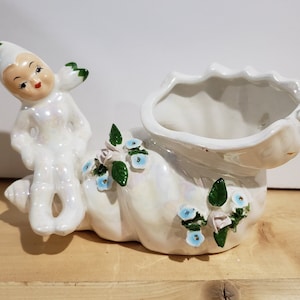 May include: A white ceramic planter shaped like a seashell with a small fairy figurine sitting on top. The planter is decorated with blue and green flowers.
