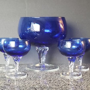 May include: A collection of cobalt blue glassware, comprising a large bowl and four smaller stemmed glasses. The bowl and glasses feature clear, twisted stems. The glassware is displayed on a speckled surface.
