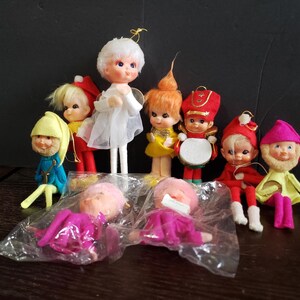May include: A collection of nine small vintage-style dolls with painted faces and felt clothing. The group includes figures dressed as an angel, a drummer, and various elves in red, blue, yellow, and pink outfits. Two dolls are sealed in plastic.