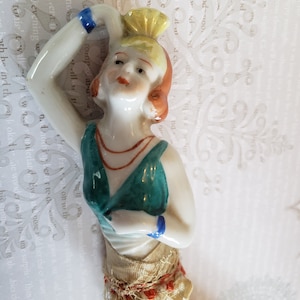 Peut inclure: Une figurine en porcelaine d'une femme vêtue d'une robe verte, d'un bandeau rouge et blanc et d'une ceinture à perles dorées et rouges. La figurine lève le bras et porte un bracelet bleu et blanc au poignet.