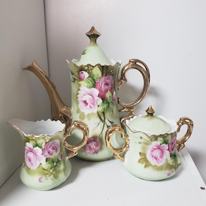 May include: A vintage porcelain tea set comprising a teapot, creamer jug, and sugar bowl. The set has a pale green base with pink rose floral designs and gold trim. The teapot is approximately 20 cm tall.