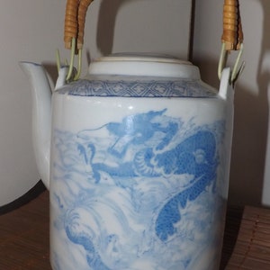 May include: White ceramic teapot with a blue dragon design. The teapot has a woven handle and a lid. The dragon is depicted in a flowing, stylized manner. The teapot is a decorative item.