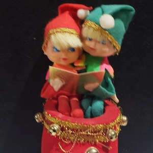 Peut inclure: Deux figurines d'elfes, l'une portant un chapeau rouge et l'autre un chapeau vert, sont assises sur un sac en velours rouge avec des garnitures dorées et des cloches. Elles tiennent un livre et chantent des chants de Noël.
