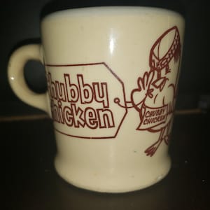 Peut inclure: Une tasse en céramique de couleur crème avec une impression graphique marron. Le motif comprend le texte "Chubby Chicken" dans un cadre rectangulaire et une illustration de dessin animé d'un poulet portant un chapeau, tenant un panneau "Chubby Chicken".