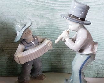 May include: A porcelain figurine set of two boys playing music. One boy is wearing a blue hat and playing a flute, while the other boy is wearing a brown hat and playing an accordion.
