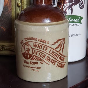 May include: A brown ceramic bottle with a cork stopper and a handle. The bottle has a label that reads "Col. Ichabod Conk's White Lightnin After Shave Tonic Bay Rum M. Hollywood 91602 7 fl. oz."
