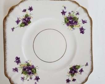 May include: A white square cake plate with a gold rim and a gold circle in the centre. The plate is decorated with purple and green violets.