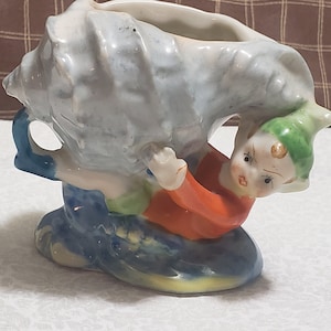 Peut inclure: Une figurine en céramique d'un lutin fantaisiste tenant un pot de fleurs en forme de coquille. Le lutin porte un chapeau vert, une chemise orange et un pantalon bleu. La coquille est peinte dans des tons de bleu et de blanc.