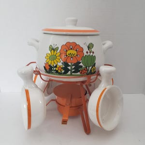 Peut inclure: Un service à fondue vintage. Le pot en céramique blanche est orné d'une bordure orange et de motifs floraux. L'ensemble comprend un support en métal, un brûleur et trois bols de service en céramique blanche avec des rayures orange.