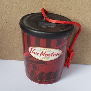 May include: A small, cylindrical Tim Hortons ornament featuring a black lid and a red body with dark red vertical stripes. The logo reads Always Tim Hortons Fresh, with the text Tim and Please Do Not Litter printed on the lower section and a red cord.