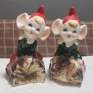 May include: Two vintage ceramic elf figurines with large ears, wearing green and red outfits, sitting on brown log-like bases.