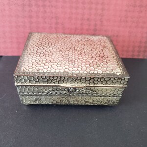 May include: A silver metal trinket box with a textured, honeycomb-like pattern on the lid. The box has a decorative floral design around the edge.