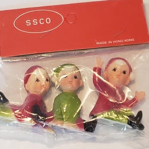 May include: Three small elf figurines in a clear plastic bag with a red cardboard header card labelled SSCO and MADE IN HONG KONG. The elves wear red and green outfits with black boots and white hats, featuring painted facial details and rosy cheeks.