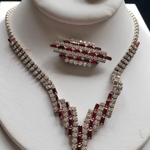 Peut inclure: Un ensemble de collier, broche et boucles d'oreilles en strass rouge et transparent. Le collier présente un design en V avec un pendentif qui pend. La broche est rectangulaire et les boucles d'oreilles sont petites et incurvées.