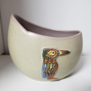 May include: A light green ceramic planter with a stylized bird design in shades of blue, green, yellow, and brown. The bird has a long beak and a large eye.