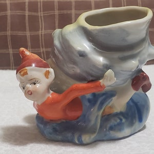 May include: A ceramic figurine of an elf holding a blue and white seashell planter. The elf is wearing a red and white hat and a red shirt. The seashell planter has a white rim and a blue and white swirl design.