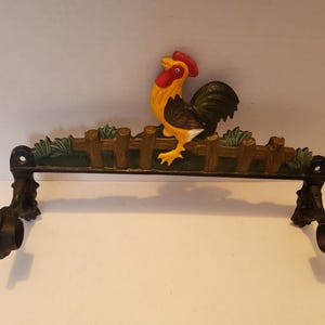 May include: A cast iron wall-mounted towel rack featuring a painted rooster perched on a brown wooden-style fence. The rooster has a red comb, yellow neck, and black tail feathers. The rack includes two black circular mounting brackets on each end.