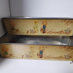 May include: Two rectangular metal planters with a light yellow background. Each planter features a charming illustration of a child in overalls, chickens, a sunflower, and a watering can. The planters have a silver trim.