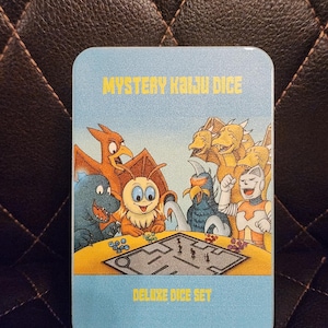 May include: A light blue metal box featuring the text "MYSTERY KAIJU DICE" and "DELUXE DICE SET". The box has a colorful illustration of various cartoon monsters playing a board game. The background is a dark brown.