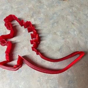 May include: Red plastic cookie cutter shaped like Godzilla, a fictional monster.