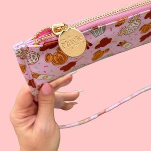 May include: Pink pencil case with a gold zipper and a gold charm that says "Sew Magic". The case is decorated with a pattern of cartoon waffles, donuts, and cupcakes. The strap is a light pink and white.