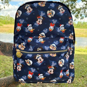 May include: A blue backpack with a pattern of Disney characters wearing sailor hats. The characters include Mickey Mouse, Minnie Mouse, Donald Duck, and Chip 'n Dale. The backpack has a gold zipper and a gold star pattern.