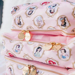 May include: Three pink cosmetic bags stacked on top of each other, featuring a repeating pattern of framed princess portraits. Each bag has a gold zipper and a gold charm with the word "Magic" on it.