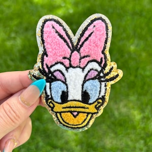 May include: Embroidered patch featuring Daisy Duck's face. The patch has a pink bow, blue eyes, and a yellow beak. The edges are outlined in black and trimmed with gold glitter. The background is a blurred green.