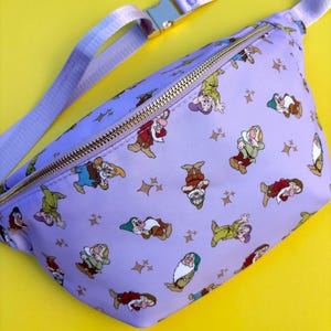 Op de afbeelding: Een paarse heuptas met een gouden rits en een patroon van de zeven dwergen uit Sneeuwwitje. De dwergen dragen hun kenmerkende outfits en zijn omringd door gouden sterren.