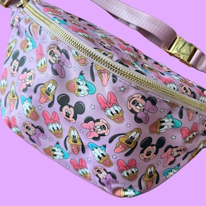 Może przedstawiać: Fioletowa saszetka biodrowa z powtarzającym się wzorem twarzy postaci z kreskówek, w tym Myszki Miki, Myszki Minnie, Kaczora Donalda i Kaczki Daisy. Torba ma złoty zamek błyskawiczny, fioletowy pasek i złotą klamrę.