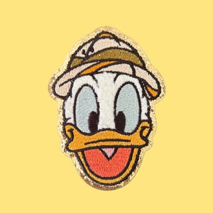 May include: Embroidered patch featuring Donald Duck's face, with a white head, blue eyes, and an orange beak. The patch has a gold glitter border and a safari hat. The background is a solid yellow.