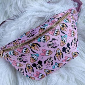 May include: Pink fanny pack with a repeating pattern of cartoon characters including Mickey Mouse, Minnie Mouse, Donald Duck, and Pluto. The bag has a gold zipper and a matching pink strap.