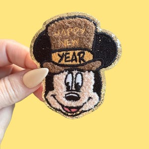Puede incluir: Un parche de Mickey Mouse con temática de Año Nuevo. El parche presenta a Mickey con un sombrero de copa marrón con las palabras "Happy New Year" bordadas. El parche tiene un borde de purpurina dorada y un fondo amarillo.