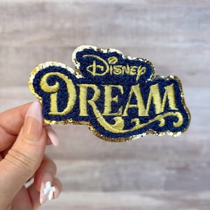 May include: A blue and gold embroidered patch with the text "Disney Dream" in a vintage font. The patch is covered in gold glitter.