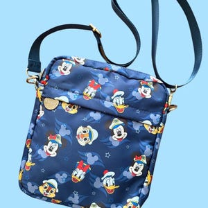 May include: Navy blue crossbody bag with a repeating pattern of Disney characters such as Mickey Mouse, Donald Duck, and Daisy Duck. The bag has a blue adjustable strap, gold-coloured hardware, and a small gold-coloured emblem.
